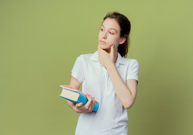 Riflessivo giovane studentessa graziosa che tiene il libro e il blocco note guardando il lato e toccando il viso isolato su sfondo verde oliva con lo spazio della copia