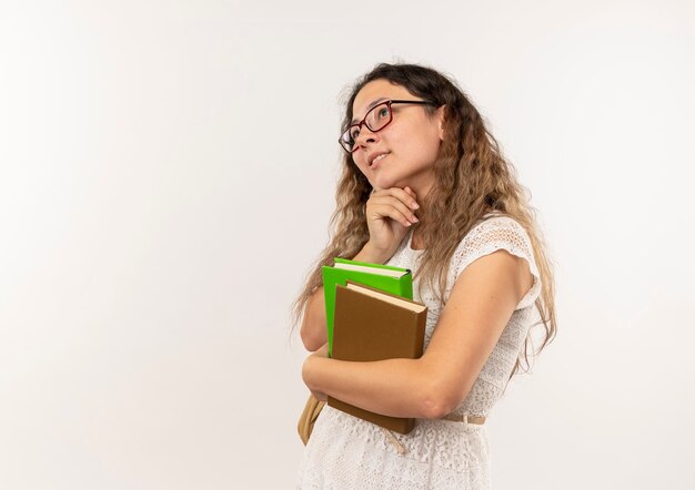 Riflessivo giovane studentessa graziosa con gli occhiali e borsa posteriore che tengono i libri mettendo la mano sotto il mento guardando il lato isolato sul muro bianco