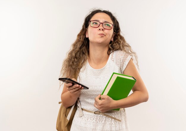 Riflessivo giovane studentessa graziosa con gli occhiali e borsa posteriore che tiene il libro e il telefono cellulare che osserva in su isolato sul muro bianco