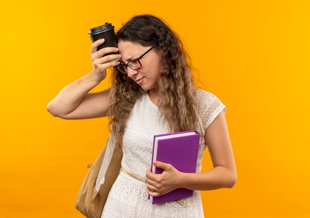 Riflessivo giovane studentessa graziosa con gli occhiali e borsa posteriore che tiene il libro guardando la testa commovente laterale con la tazza di caffè di plastica isolata sulla parete gialla