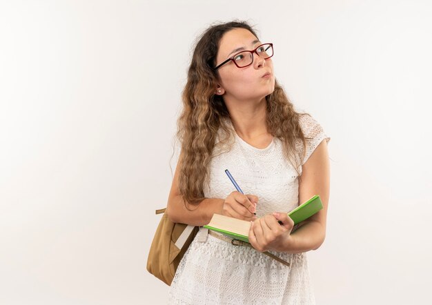 Riflessivo giovane studentessa graziosa con gli occhiali e borsa posteriore scrivendo sul libro guardando il lato isolato sul muro bianco