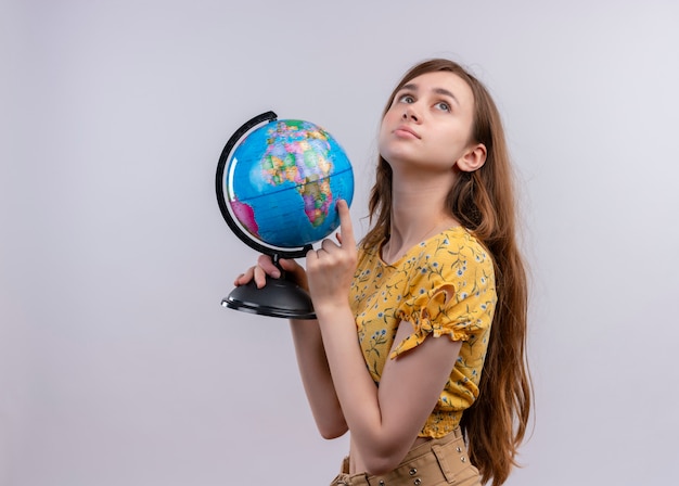 Riflessivo ragazza giovane azienda globo mettendo il dito su di esso e guardando in alto sulla parete bianca isolata con lo spazio della copia