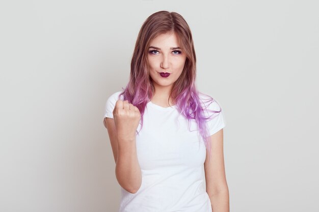Rigorosa femmina seria con i capelli lilla che indossa la maglietta bianca che mostra il pugno con espressione arrabbiata, avvertendo di fare cose cattive, in posa isolata sopra il muro grigio