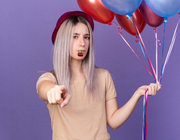 Rigorosa giovane bella ragazza che indossa un cappello da festa che tiene palloncini che soffiano fischio di festa che ti mostra gesto isolato sulla parete blu