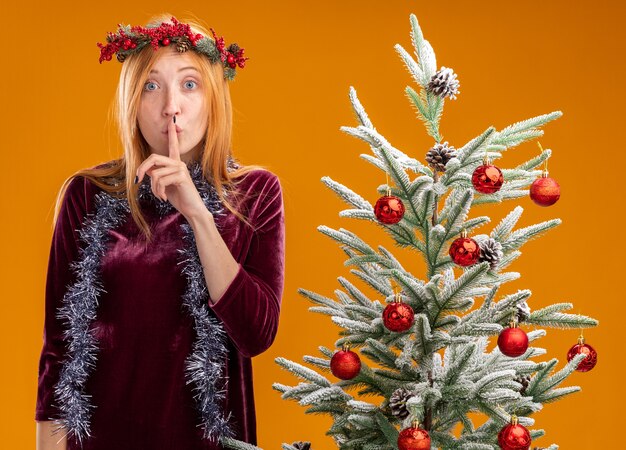 Rigorosa giovane bella ragazza in piedi vicino all'albero di Natale che indossa abito rosso e corona con ghirlanda sul collo che mostra gesto di silenzio isolato su sfondo arancione