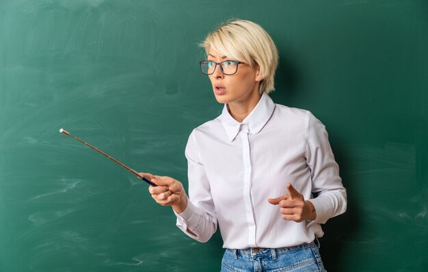 Rigorosa giovane bionda insegnante di sesso femminile con gli occhiali in aula in piedi di fronte alla lavagna guardando e puntando a lato con puntatore stick e dito