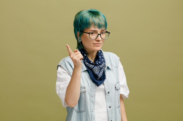 Rigorosa giovane donna che indossa occhiali bandana sul collo guardando la fotocamera che mostra il dito indice isolato su sfondo verde oliva