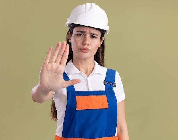 Rigorosa giovane donna del costruttore in uniforme che mostra il gesto di arresto isolato sulla parete verde oliva