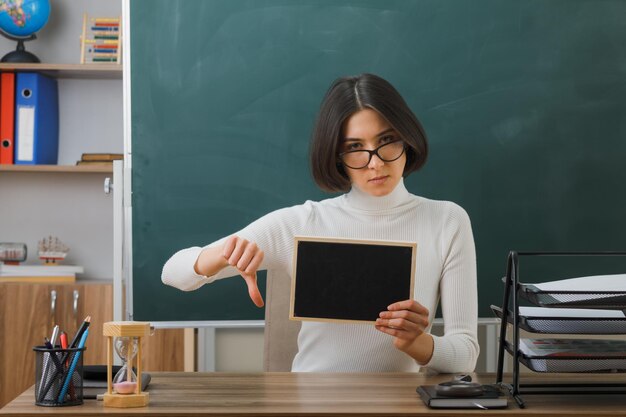 rigoroso che mostra i pollici verso il basso giovane insegnante femminile che indossa occhiali con in mano una mini lavagna seduta alla scrivania con gli strumenti della scuola in classe