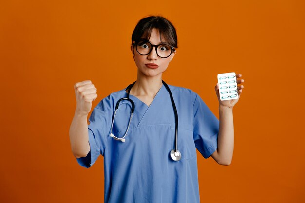 Rigoroso che mostra il pugno che tiene le pillole giovane dottoressa che indossa uno stetoscopio fith uniforme isolato su sfondo arancione