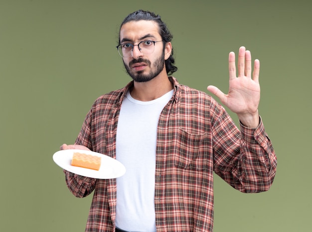 Rigoroso giovane bel ragazzo di pulizia che indossa t-shirt tenendo la spugna sul piatto che mostra il gesto di arresto isolato sulla parete verde oliva