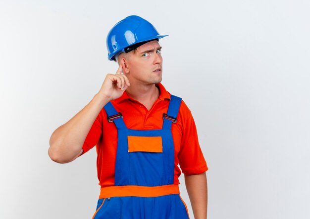 Rigoroso giovane costruttore maschio che indossa l'uniforme e casco di sicurezza che mette il dito sull'orecchio su bianco