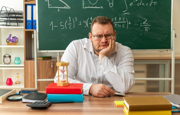 rigoroso giovane insegnante con gli occhiali seduto alla scrivania con materiale scolastico in classe tenendo la mano sul mento guardando davanti