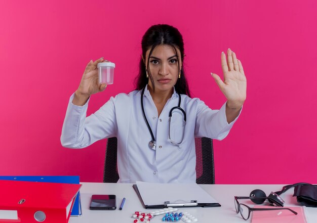 Rigoroso giovane medico femminile che indossa abito medico e stetoscopio seduto alla scrivania con strumenti medici che tengono bicchiere medico facendo gesto di arresto isolato sulla parete rosa
