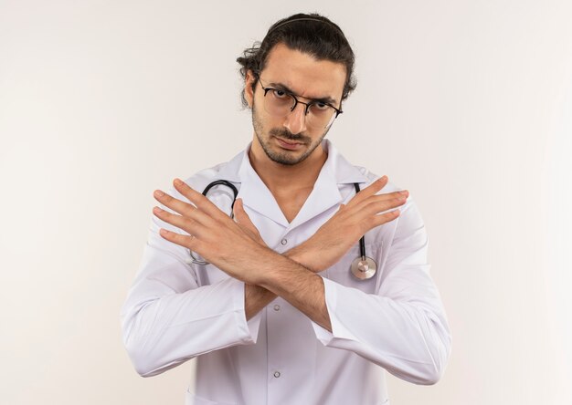 Rigoroso giovane medico maschio con vetri ottici che indossa una veste bianca con lo stetoscopio che mostra il gesto di no sulla parete bianca isolata con lo spazio della copia