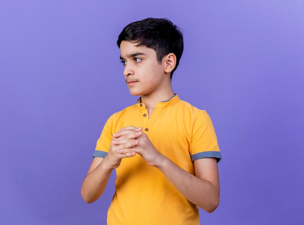 Rigoroso giovane ragazzo caucasico guardando a lato tenendo le mani insieme isolato su sfondo viola con copia spazio