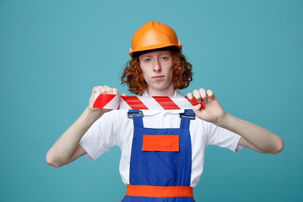 Rigoroso guardando la fotocamera giovane costruttore uomo in uniforme tenendo il nastro adesivo isolato su sfondo blu