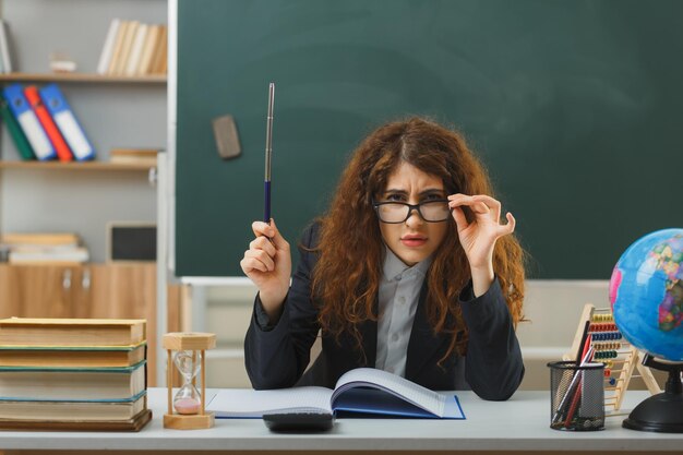 rigoroso guardando la fotocamera giovane insegnante femminile che indossa occhiali tenendo il puntatore seduto alla scrivania con strumenti scolastici in classe
