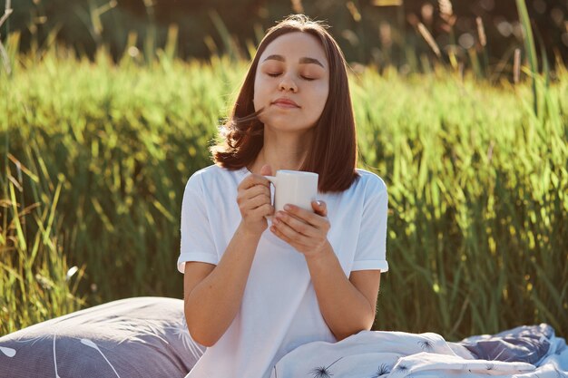 Rilassata donna dai capelli scuri che indossa una maglietta bianca in stile casual in posa all'aperto con una tazza di tè caldo o caffè