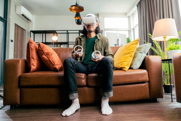 Rilassati casual uomo adulto asiatico che indossa le cuffie per realtà virtuale e tiene i controller Gioca in uno sport Videogioco a casa Gioca a VR gioco di sport attivo online nel soggiorno