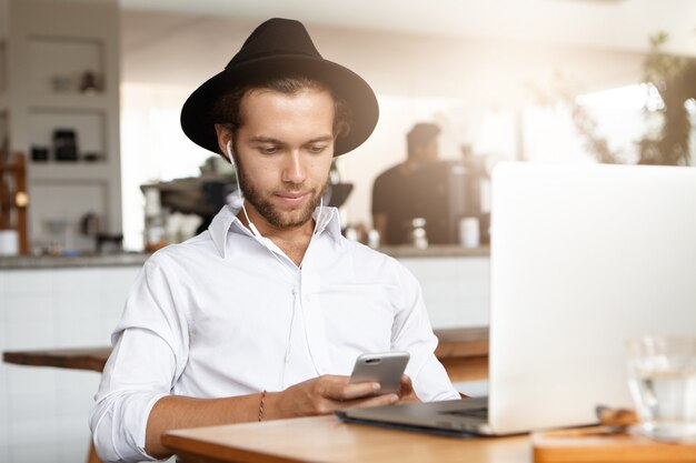 Rimanere in contatto. Bel giovane utilizzando smartphone, seduto al bar davanti al computer portatile in auricolari in attesa del suo pranzo