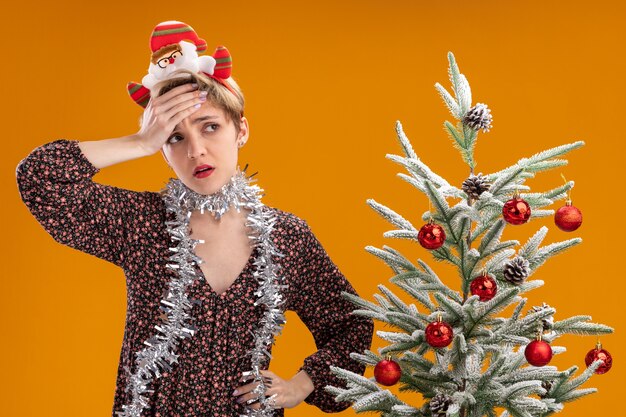 rimpiangendo la giovane ragazza carina che indossa la fascia di babbo natale e la ghirlanda di orpelli intorno al collo in piedi vicino all'albero di natale decorato tenendo la mano sulla fronte e sulla vita guardando il lato isolato sul muro arancione