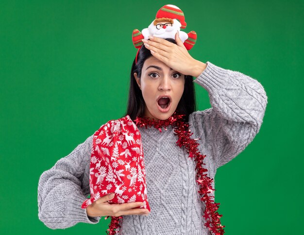Rimpiangendo la giovane ragazza caucasica che indossa la fascia di Babbo Natale e la ghirlanda di orpelli intorno al collo tenendo il sacco del regalo di Natale tenendo la mano sulla fronte isolata sulla parete verde