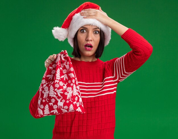 Rimpiangendo la ragazza che indossa il cappello della Santa che tiene il sacco del regalo di Natale che guarda l'obbiettivo tenendo la mano sulla testa isolata su fondo verde