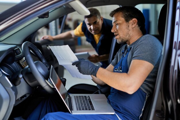 Riparatore automobilistico che parla con il suo collega mentre esegue la diagnostica dell'auto e analizza i dati in un'officina