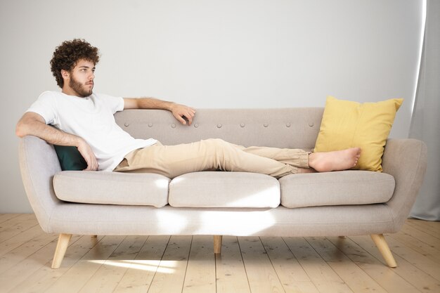 Riposo, relax e concetto di svago. Giovane attraente con stoppia e capelli voluminosi sdraiato comodamente sul divano grigio in soggiorno e guardare la tv, godersi la partita di calcio o una serie