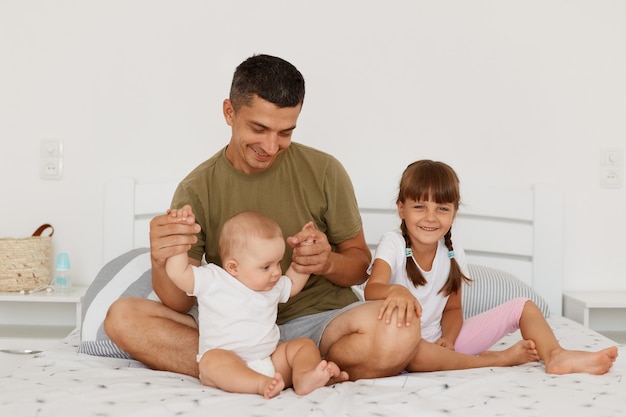 Ripresa in interni di una famiglia felice e positiva in posa in camera da letto luminosa, giovane padre adulto seduto sul letto con figlie piccole, bambine in età prescolare, genitorialità.