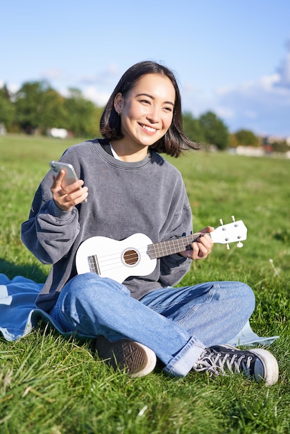 Ripresa verticale di una ragazza coreana sorridente seduta nel parco con ukulele e smartphone che utilizza l'app mobile per