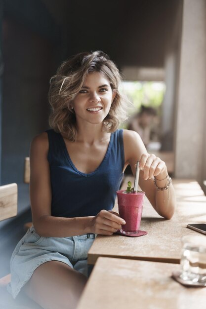 Ripresa verticale elegante studentessa prendersi una pausa bere frullato sedersi finestra bar caffetteria interna sorridente felice fotocamera parlando avere una piacevole conversazione spensierata dopo le lezioni universitarie