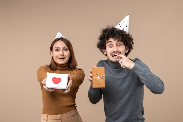 Riprese di nuovo anno con la giovane coppia indossa la ragazza felice del cappello del nuovo anno con il cuore e il ragazzo sorpreso con il regalo su gray