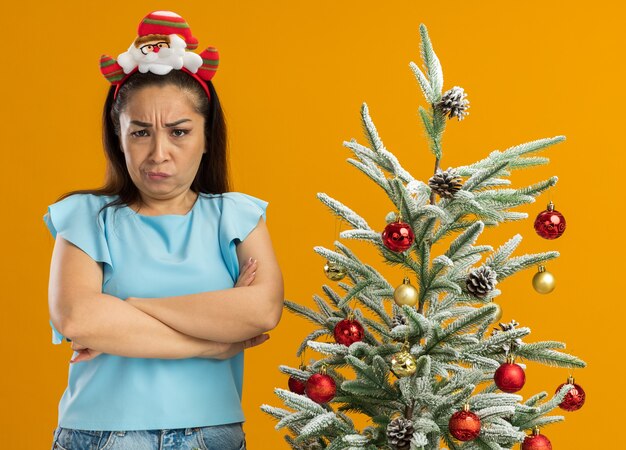 Risentita giovane donna in blu in alto indossando divertenti natale cerchio sulla testa guardando la fotocamera con la fronte accigliata con le braccia incrociate in piedi accanto a un albero di natale su sfondo arancione