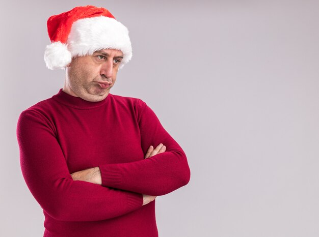 Risentito uomo di mezza età che indossa il cappello di Babbo Natale che guarda da parte con le braccia incrociate in piedi su un muro bianco