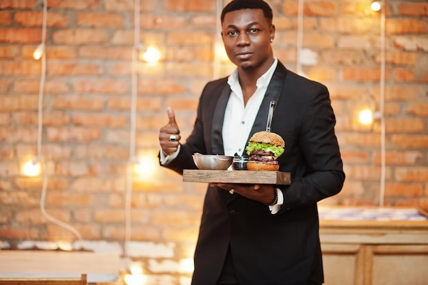 Rispettabile giovane afroamericano in abito nero tenere vassoio con doppio hamburger e mostrare il pollice contro il muro di mattoni del ristorante con luci