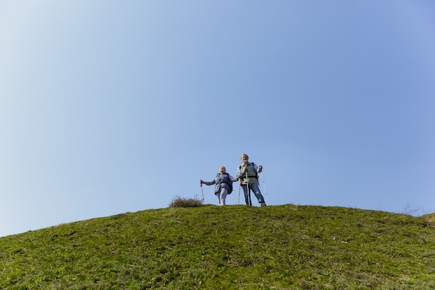 Risultati comuni. Coppia di famiglia invecchiato dell'uomo e della donna in abito turistico che cammina al prato verde vicino agli alberi in una giornata di sole. Concetto di turismo, stile di vita sano, relax e solidarietà.