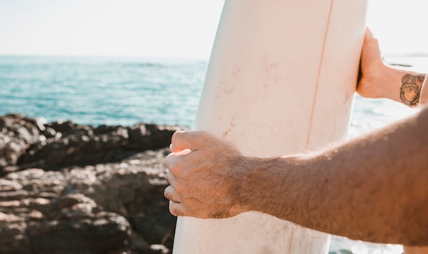 Ritaglia le mani tenendo la tavola da surf vicino a pietre e mare