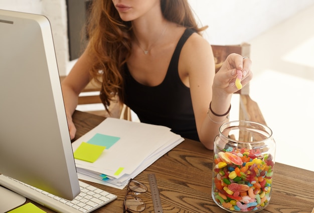 Ritagliata colpo dell'impiegato femminile caucasico che prende la marmellata dal barattolo grande al lavoro. La ragazza mangia caramelle per miglioramento del lavoro di un cervello durante un duro lavoro sopra il rendiconto finanziario