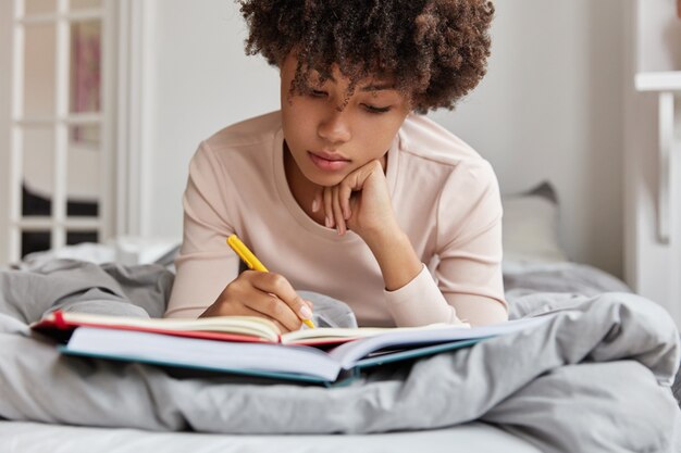 Ritagliata colpo di donna dalla pelle scura dall'aspetto piacevole scrive note nel taccuino dal libro, ha concentrato lo sguardo verso il basso, si trova sul letto comodo