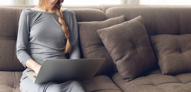 Ritagliata colpo di giovane donna bionda con la treccia che lavora su un laptop seduto sul comodo divano scuro a casa, luce calda retroilluminata. Freelance o concetto di stile di vita.