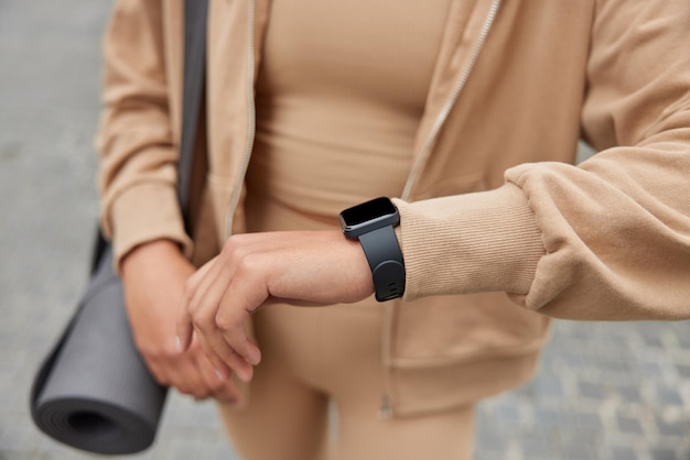 Ritagliata colpo di giovane donna sportiva senza volto controlla i risultati di fitness su smartwatch moderno utilizza l'applicazione necessaria trasporta karemat arrotolato vestito in pose di abbigliamento sportivo su sfondo sfocato