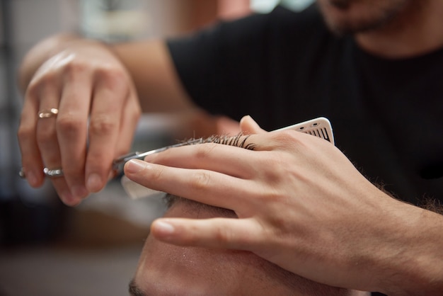 Ritagliata ravvicinata di un barbiere professionista con forbici e pettine mentre dava un taglio di capelli al suo cliente.