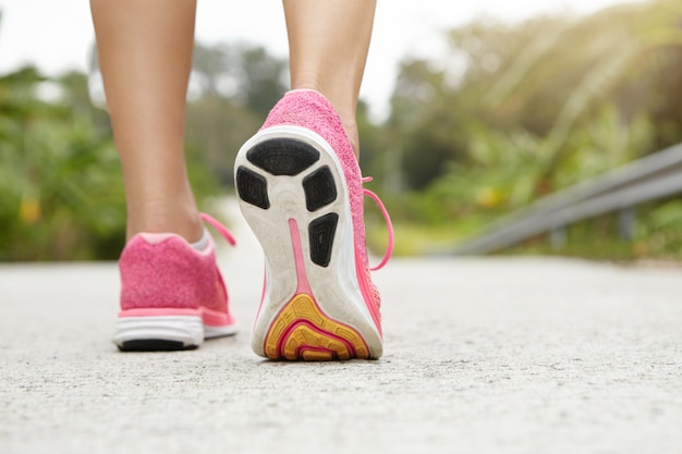 Ritagliata ripresa posteriore della ragazza atletica che indossa scarpe da ginnastica rosa durante le escursioni o fare jogging sul marciapiede all'aperto. Pareggiatore donna con belle gambe in forma facendo allenamento.