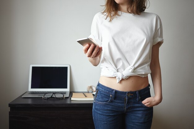 Ritagliata ritratto di moda giovane donna europea in jeans e t-shirt utilizzando il cellulare al chiuso, scaricando la nuova applicazione, in piedi al muro bianco con computer portatile aperto con schermo vuoto