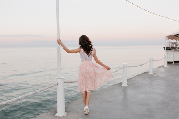 Ritratto a figura intera dal retro dell'elegante ragazza bruna che cammina lungo il molo dell'oceano e gode di una splendida vista mattutina