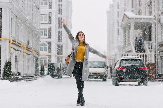 Ritratto a figura intera del modello femminile ispirato in cappotto alla moda in posa con piacere nella città invernale. Foto all'aperto di donna bionda felice che gode della nevicata durante la passeggiata intorno alla città.