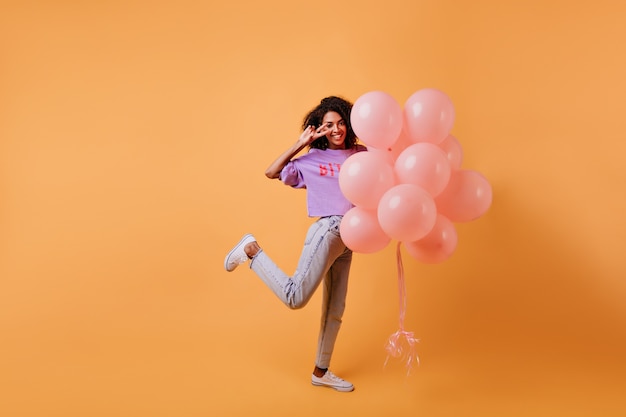 Ritratto a figura intera della ragazza africana ispirata in piedi su una gamba con palloncini. Bella signora di buon umore che festeggia il compleanno.