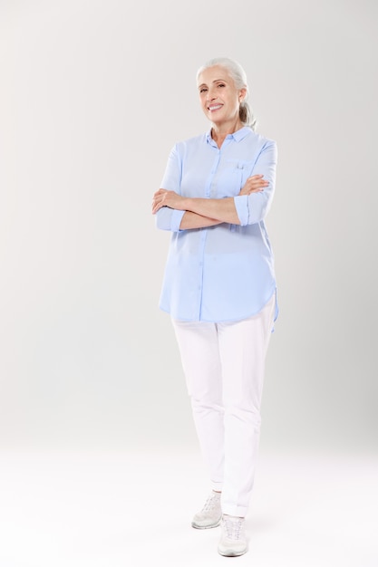 Ritratto a figura intera di affascinante vecchia signora in camicia blu e pantaloni bianchi, in piedi con le mani incrociate